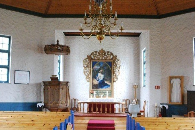 Interiøret i Digermulen kirke med et alter dominert av et stort maleri innrammet av dekorative utskjæringer. Foran alteret er det en rød alterring og en trapp med rød løper. Til venstre for alteret er en utsmykket prekestol, og en lysekrone henger fra taket. De teksturerte hvite veggene og blå benkerader står i kontrast til det lyse tregulvet. Foto.