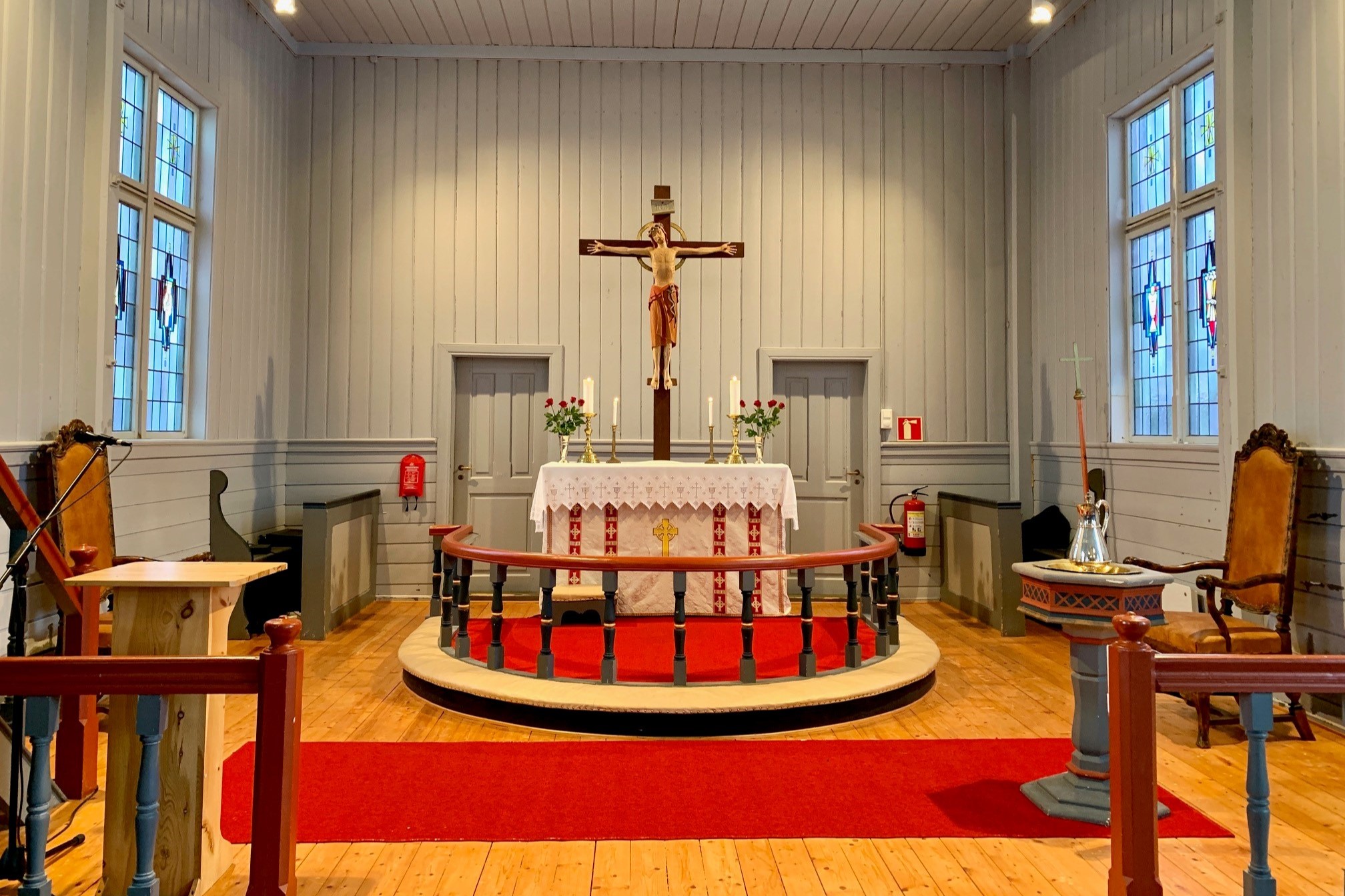 Alteret i Gimsøy kirke med et sentralt krusifiks, flankert av lysestaker og roser på et hvitt alterbord med røde detaljer. En rød løper leder opp til alterringen. Fargede glassvinduer på hver side slipper inn mykt lys som fremhever de hvitmalte treveggene og det lyse tregulvet. Foto.