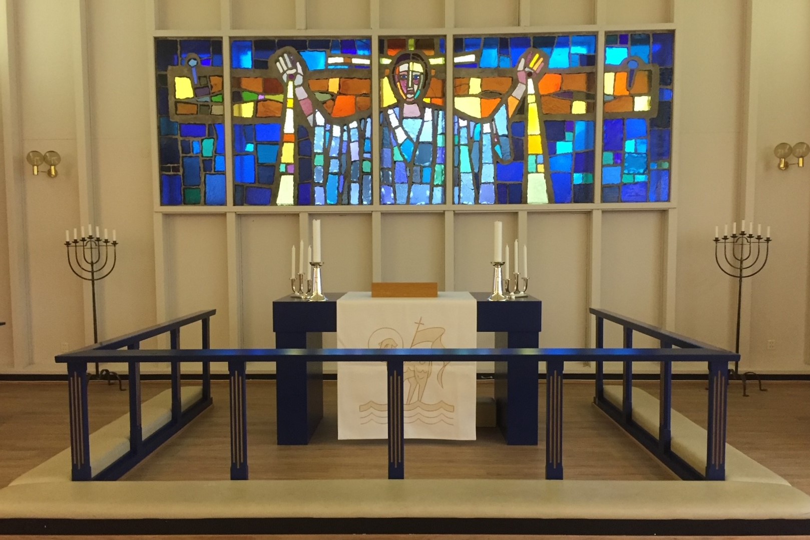 Alteret i Henningsvær kirke med et fargerikt glassmaleri som viser Jesus med utstrakte armer i bakgrunnen. Alterbordet er dekorert med hvite lysestaker og en duk med et religiøst symbol. Alterringen og rekkverket rundt alteret er malt i blått, noe som skaper en harmonisk og fredelig atmosfære i kirkerommet. Foto.