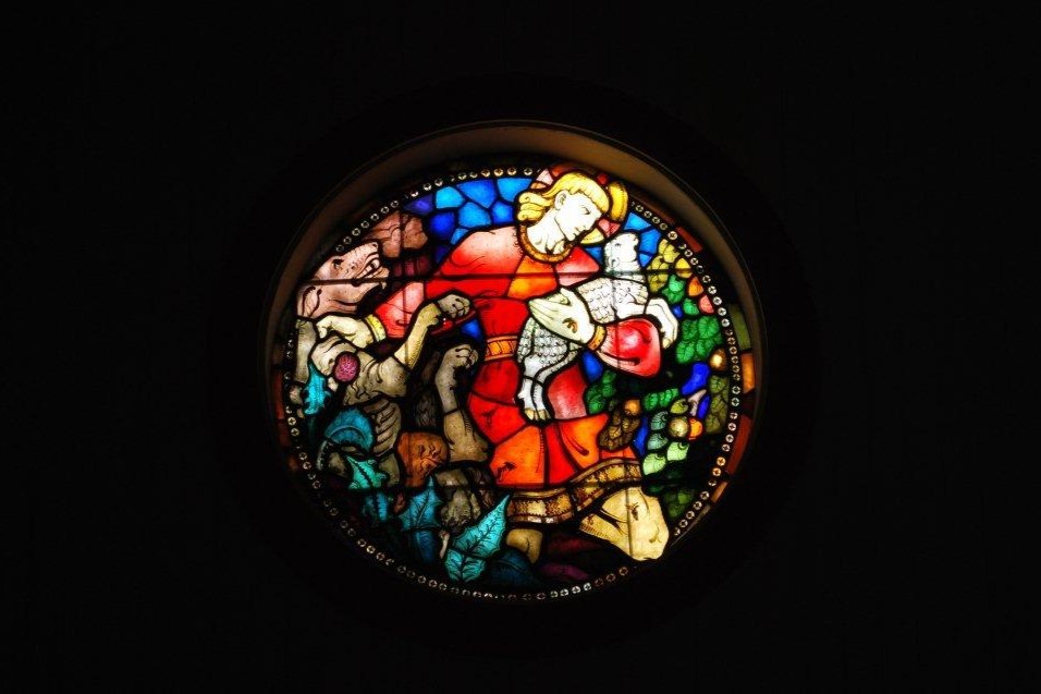 Et rundt glassmaleri i Strandlandet kirke som viser en fargerik scene med Jesus som bærer et lam. Maleriet er rikt dekorert med detaljerte figurer og levende farger som rød, blå, grønn og gul, som skaper en varm og inspirerende atmosfære. Foto.