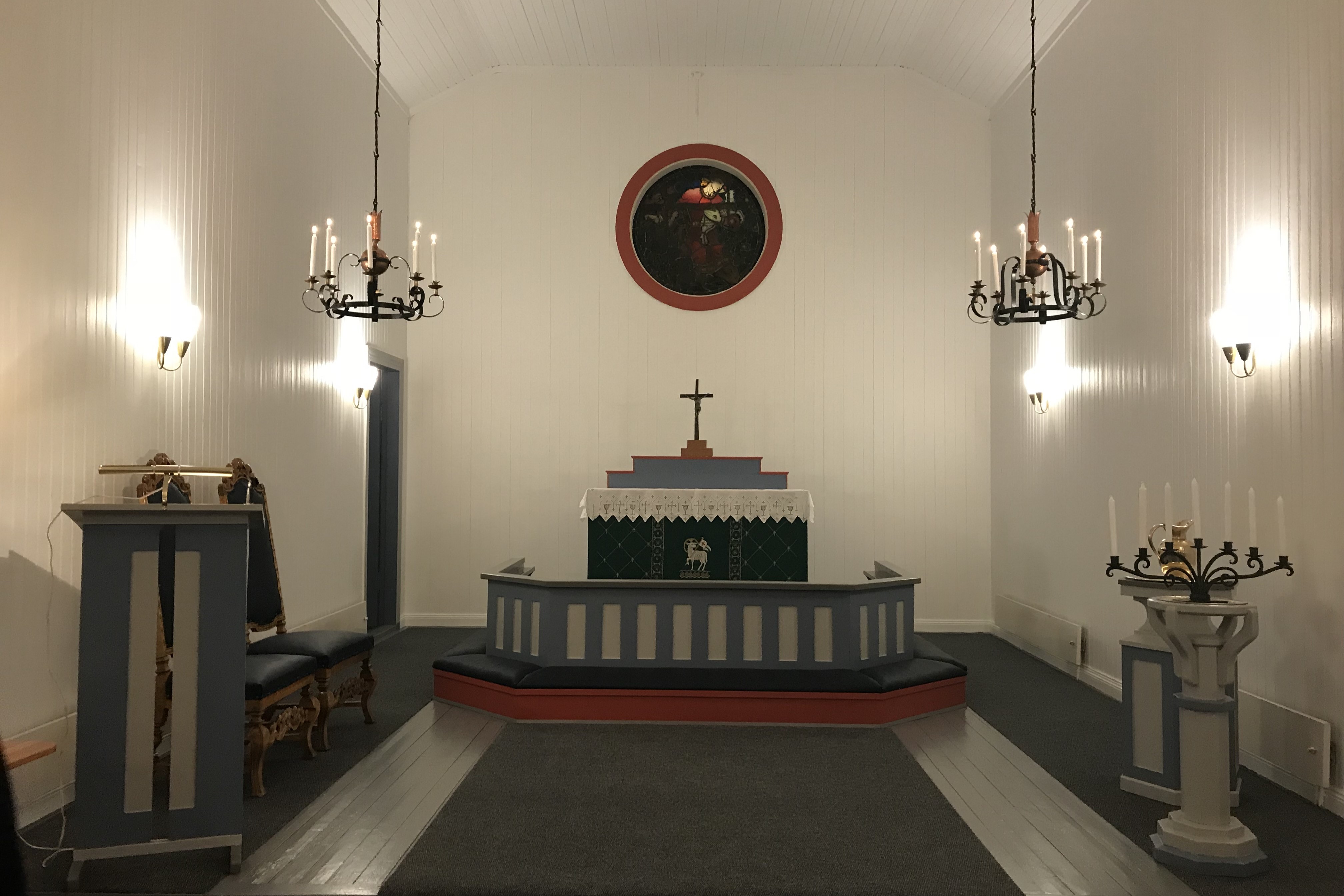 Alteret i Strandlandet kirke med et krusifiks på et enkelt alterbord dekket med en hvit duk. Alterringen er malt i blått og hvitt, og bak alteret henger et rundt glassmaleri. Rommet er opplyst av lysekroner og vegglamper, som tilfører varme til de hvitmalte veggene. Prekestolen til venstre er malt i blått og hvitt, og en kandelaber med lys står til høyre. Foto