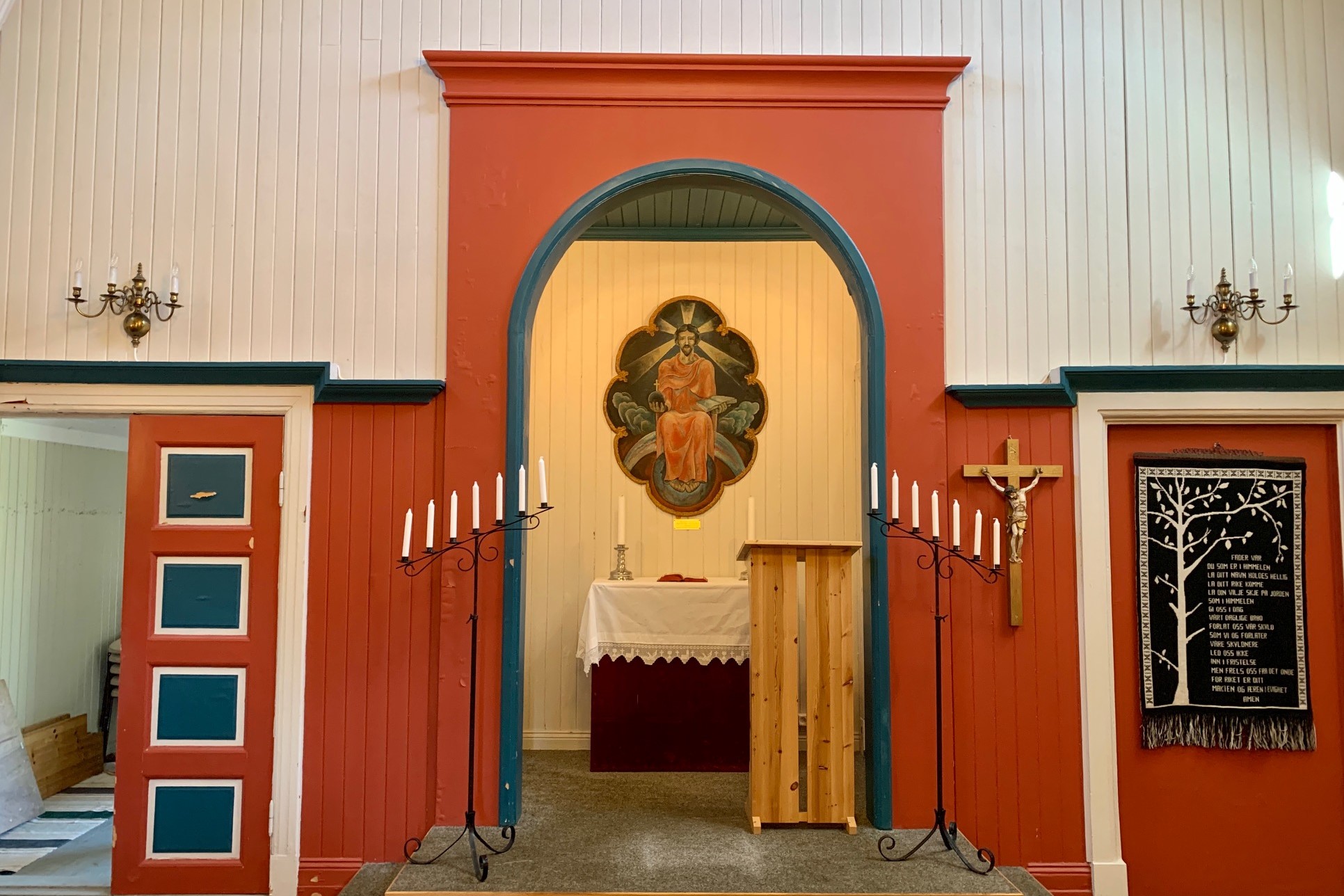 Interiøret i Svolvær kapell viser et enkelt alter med et malt bilde av Jesus i midten. De røde og blå detaljene rundt alteret gir rommet en varm og innbydende atmosfære. På hver side av alteret står smijerns staker med hvite lys, og til høyre henger et krusifiks og et veggteppe med en bønn. De hvitmalte veggene og taket skaper en lys og åpen følelse i rommet.