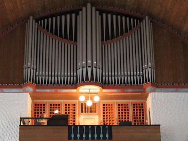 Orgelet i Svolvær kirke er et imponerende instrument som dominerer galleriet med sine mange piper og detaljrike utforming. De vertikale pipene strekker seg opp mot taket, mens den varme treinnrammingen skaper en harmonisk kontrast til de lyse pipene. Under orgelet henger en enkel lysekrone som tilfører en myk belysning til området, og gir en følelse av ro og andakt. Foto.