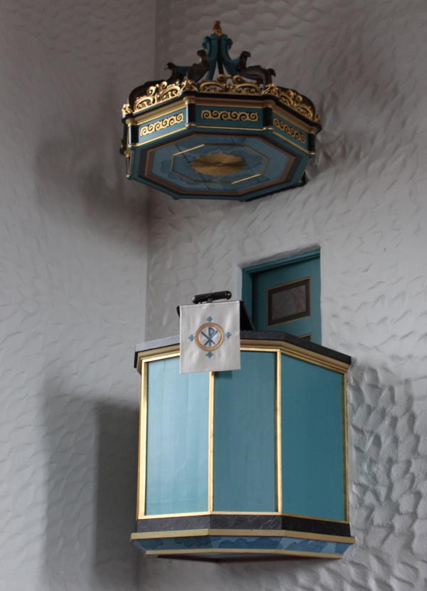 Prekestolen i Svolvær kirke er en vakker og detaljert struktur, malt i blått og dekorert med gullkanter. Over prekestolen henger en utsmykket lydhimmel, også i blått og gull, som tilfører en elegant touch. En hvit duk med et religiøst symbol pryder fronten av prekestolen, som står mot en teksturert hvit vegg, og skaper en kontrast som fremhever dens skjønnhet og betydning i kirkerommet. Foto.