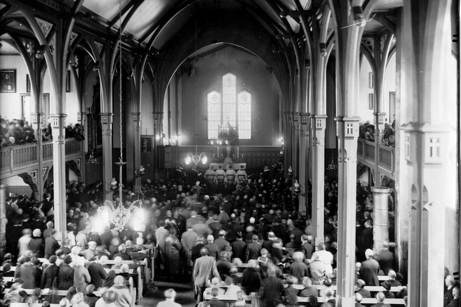 Vågan kirke, kjent som Lofotkatedralen, under presteordinasjonen 9. mai 1929. Kirken er fylt til randen av menighetsmedlemmer, og atmosfæren er høytidelig. De gotiske søylene og buene strekker seg mot taket, mens lyset fra de store glassvinduene i bakgrunnen skaper en stemningsfull belysning. Dette sort-hvitt-bildet fanger et historisk øyeblikk i kirkens liv. Foto.