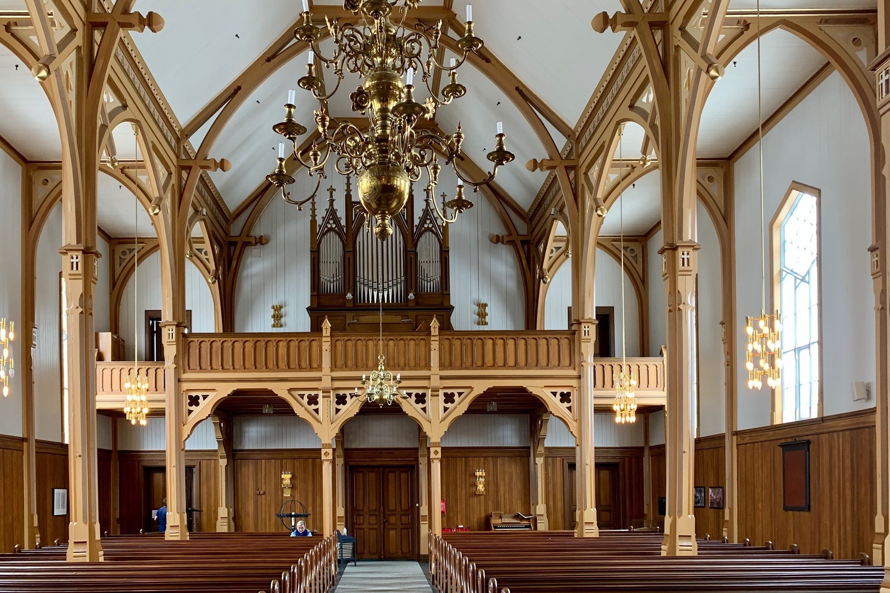 Interiøret i Vågan kirke, også kjent som Lofotkatedralen, med gotiske søyler, buer og en stor lysekrone. Orgelgalleriet i bakgrunnen tilfører en følelse av storhet, mens varme trefarger og detaljer skaper en harmonisk atmosfære. Foto.