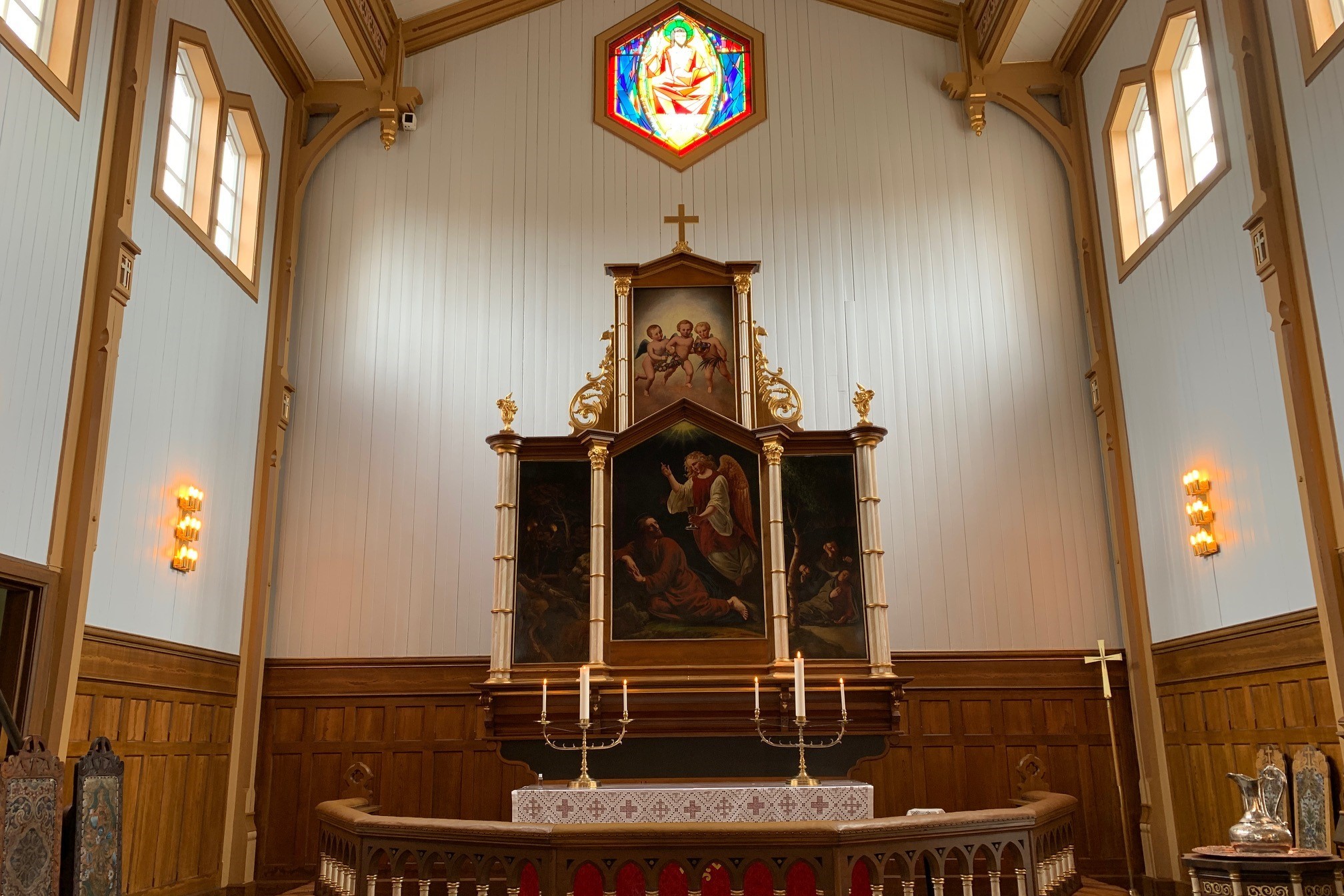 Alteret i Vågan kirke med et stort sentralt maleri, flankert av mindre bilder og gyldne dekorasjoner. Over alteret henger et fargerikt glassmaleri, og høye vinduer slipper inn naturlig lys. Foto.