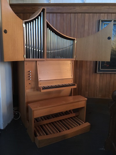 Nærbilde av kororgelet i Vågan kirke, med åpne tredeksler som viser orgelets piper. Spillepulten har en rad med tangenter og et pedalbrett nederst. Orgelet er laget av lyst tre og står mot en bakgrunn av matchende trepaneler, som skaper en harmonisk og klassisk estetikk. Foto.