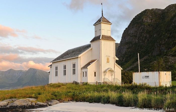 Gimsøy kirke, en hvitmalt trekirke med firkantet klokketårn, ligger i et åpent landskap ved stranden med fjell i bakgrunnen og en blå himmel med rosa skyer. Foto av Per Arvid Åsen.