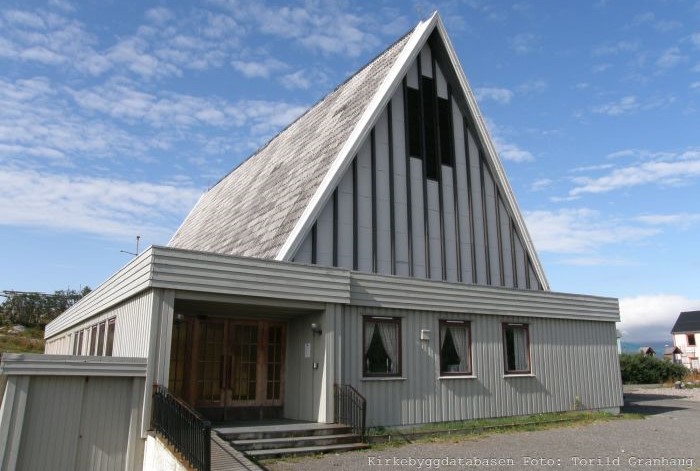 Henningsvær kirke, en moderne, grå kirke med et bratt, trekantet tak og store vinduer i fronten, ligger under en blå himmel med lette skyer. Foto av Torild Granhaug.