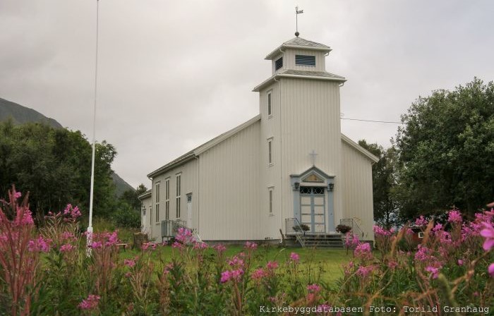Strandlandet kirke, en hvitmalt trekirke med et firkantet klokketårn og blå detaljer, omgitt av blomster og trær. Foto av Torild Granhaug.