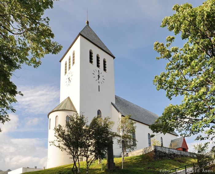 Svolvær kirke, en hvitmalt bygning med et klokketårn med tre klokkerader og en klokke. Kirken har et skråtak og står omgitt av trær. Foto av Per Arvid Åsen.