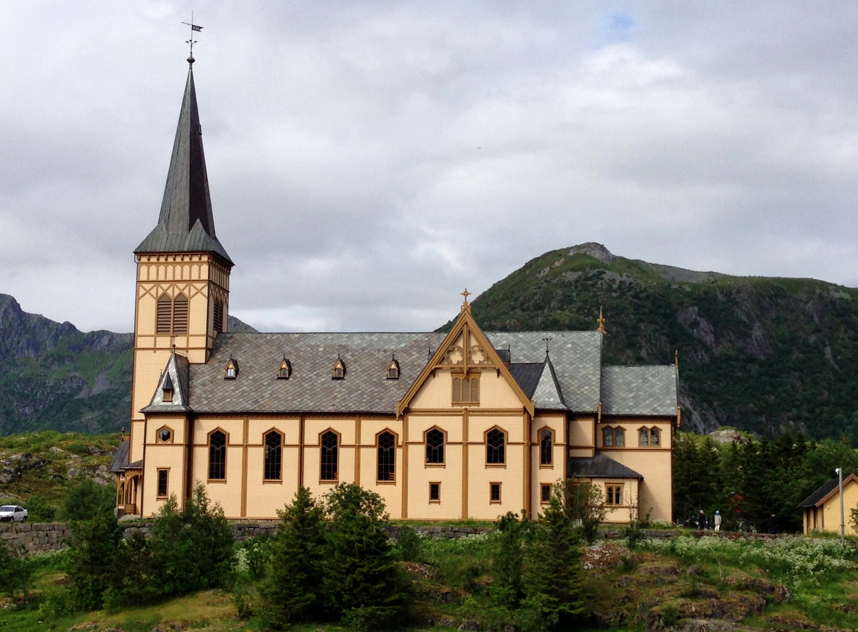 Lofotkatedralen, en stor, gul trekirke med høyt spir og gotiske vinduer, ligger i et grønt landskap med fjell i bakgrunnen. Foto.