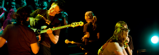 Et band opptrer på en scene, med en bassist i forgrunnen som spiller intenst. Flere personer, inkludert en sanger med mikrofon, er synlige på scenen. Bakgrunnen er mørk med fargerike lys som skaper en livlig atmosfære. Foto.